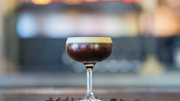 A glass of dark liquid on a reflective surface with scattered coffee beans around it.