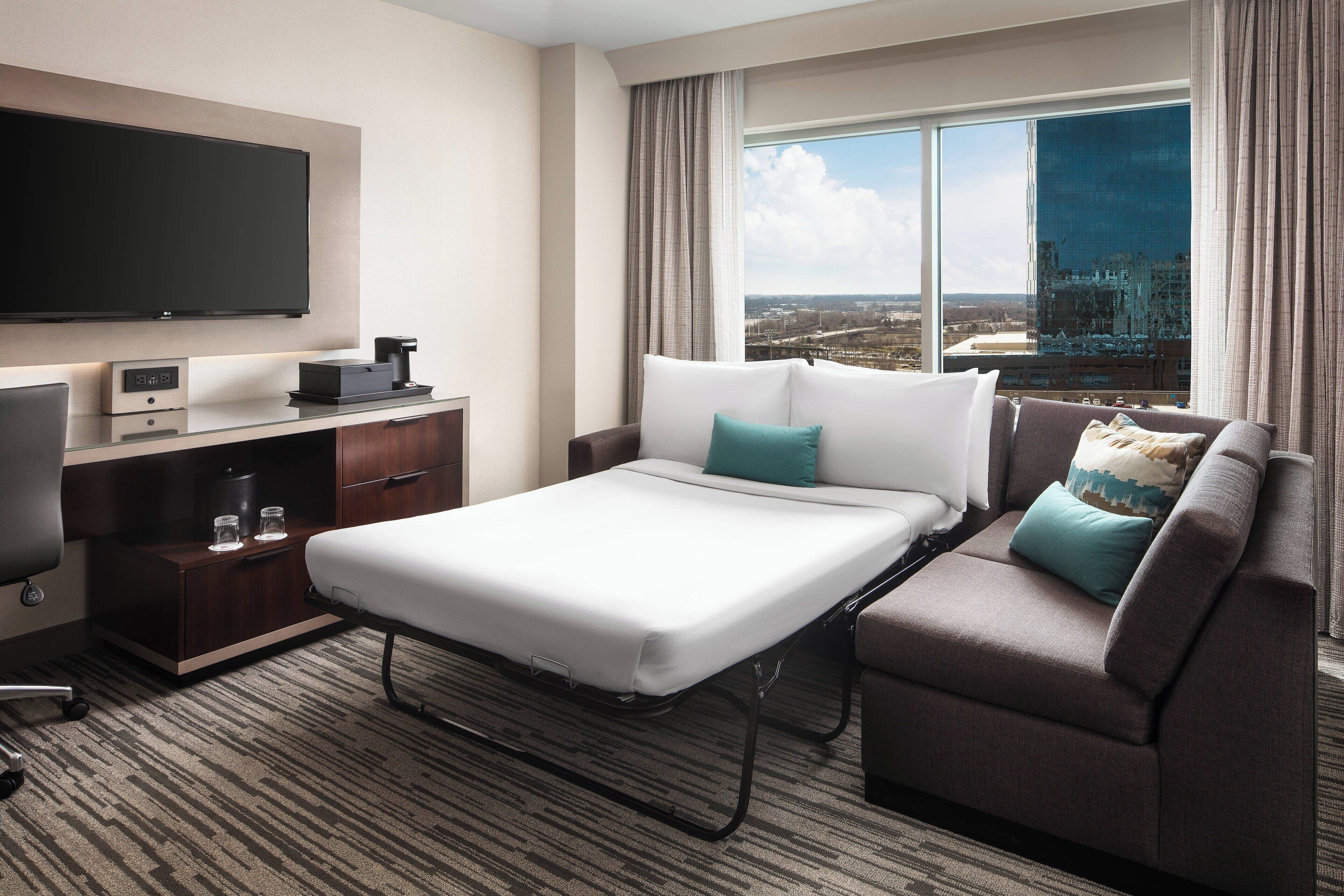 A hotel room with a bed, a sofa, a TV, a desk with a computer, and a window with a city view.
