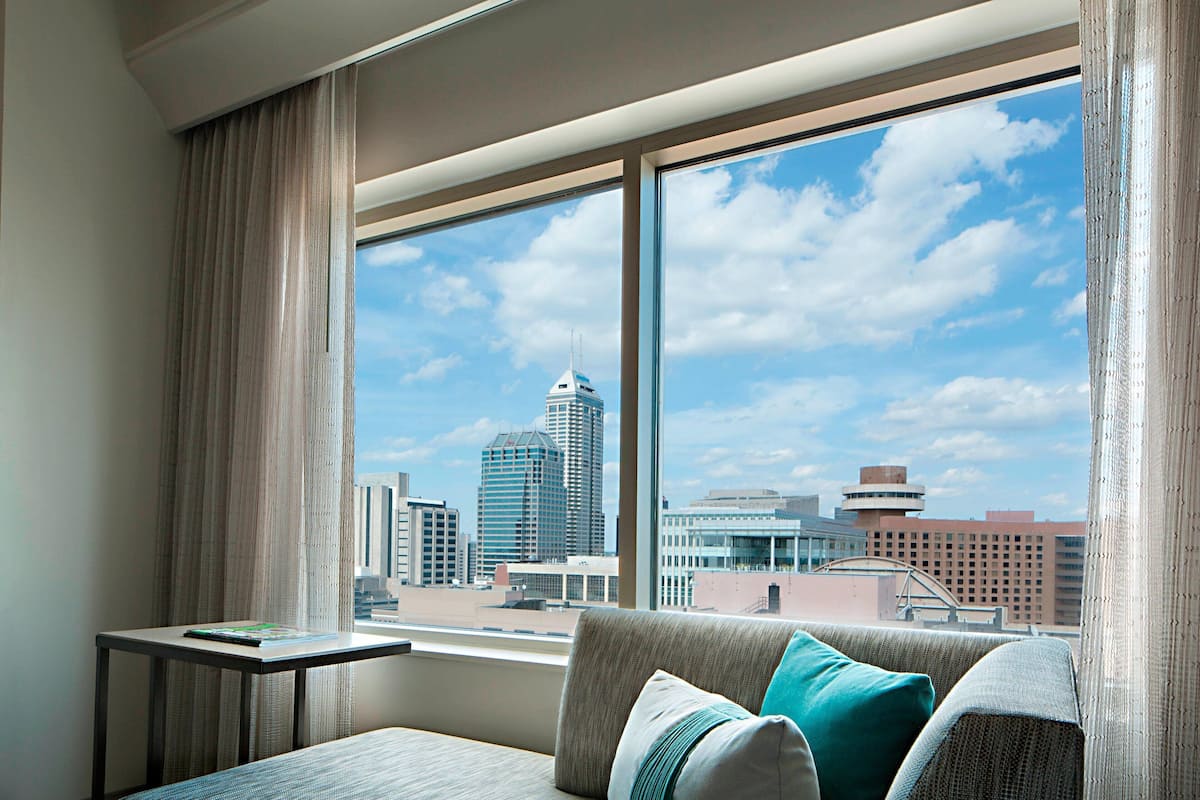 Habitación de hotel con un ventanal grande que ofrece vistas a la ciudad, un sofá con cojines y una mesita con un libro.