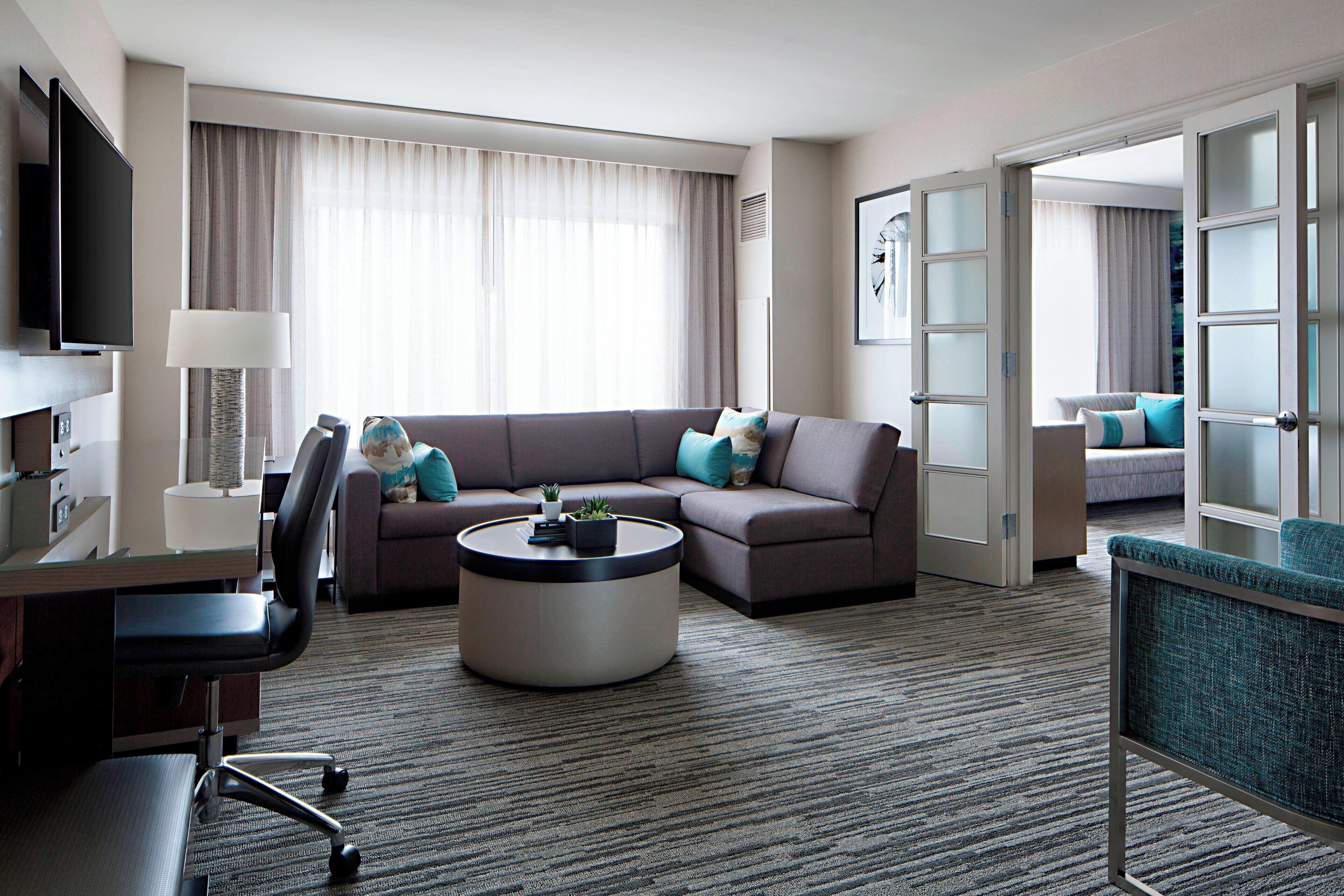 Une chambre d’hôtel moderne avec un canapé, une table basse ronde, un bureau avec une chaise, un téléviseur et un lit en arrière-plan.