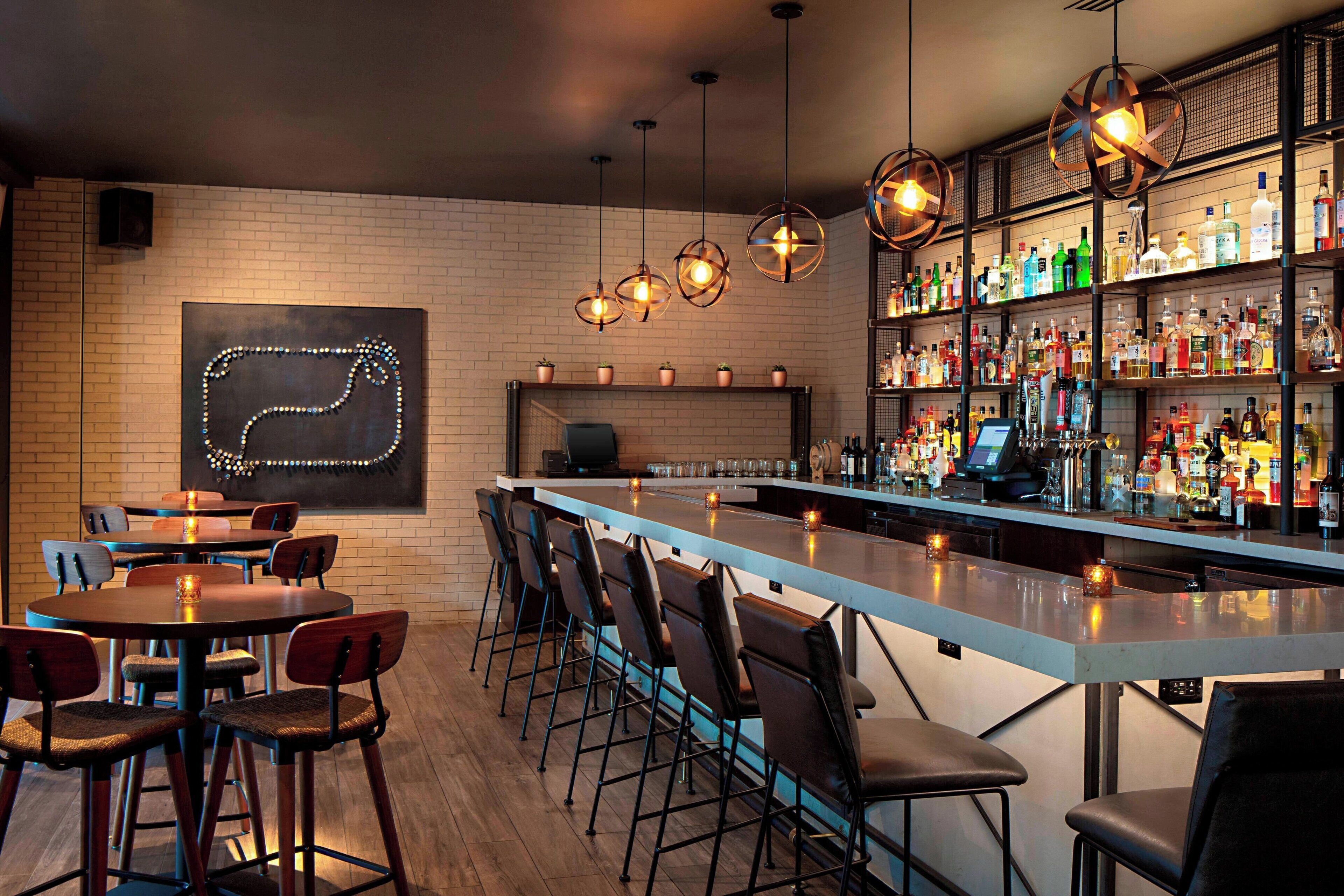 Una zona de bar con una barra alargada, varios taburetes y una pared de ladrillo con una lámpara decorativa.