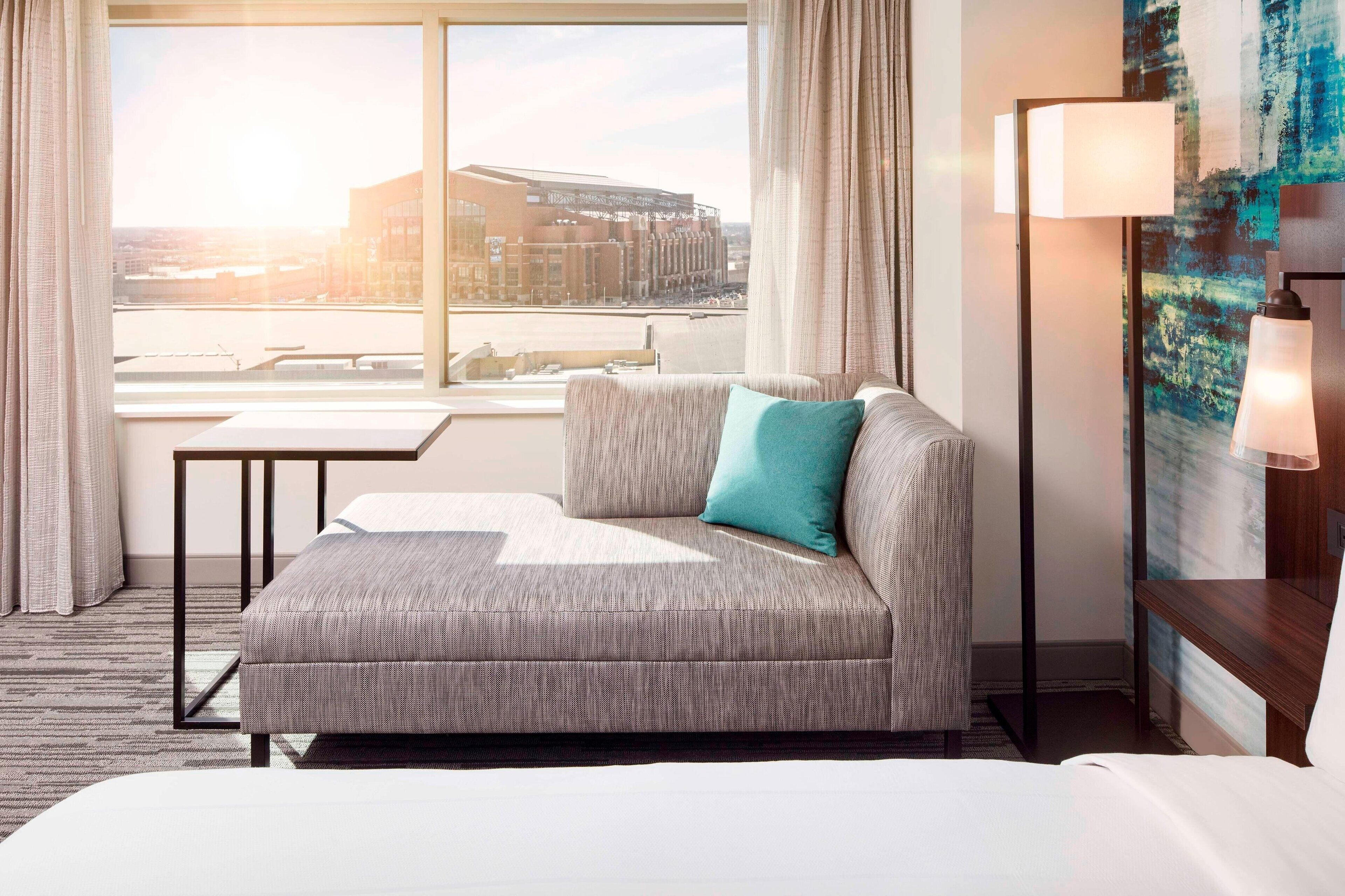 A modern hotel room with a sofa, a small table, a lamp, and a large window offering a city view.