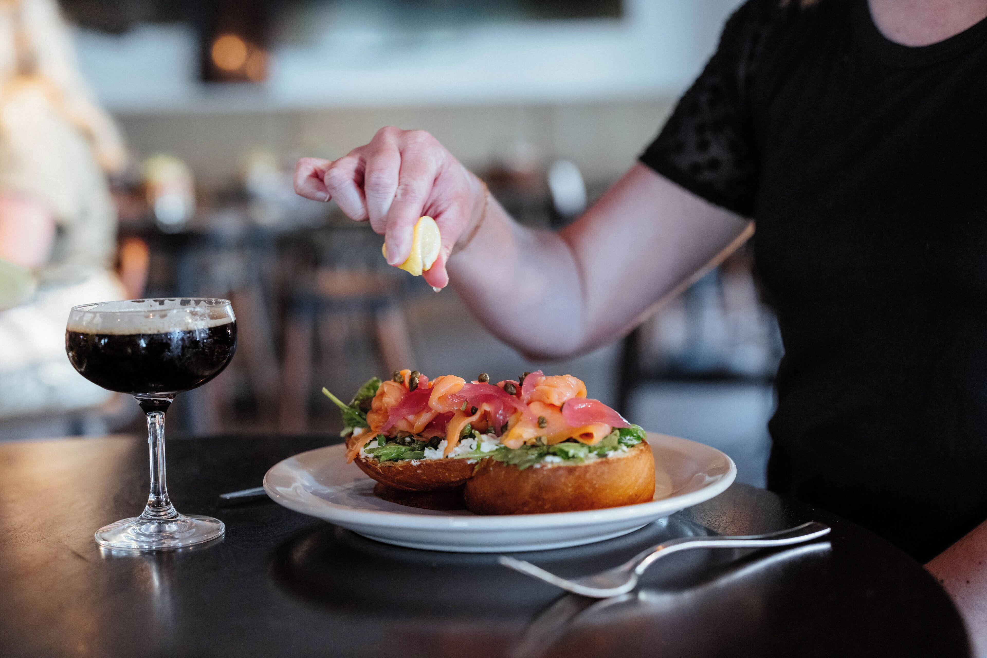 A person holding a lemon over a plate with a sandwich and a glass of dark beverage.