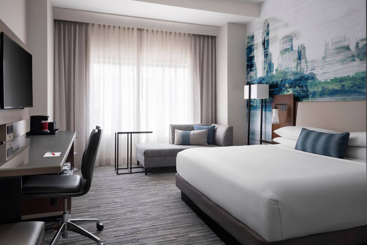 Una habitación de hotel con cama, escritorio, silla, televisor, ventana con cortinas y un cuadro en la pared.