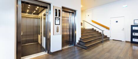 Un moderno ascensor con pantalla digital, una escalera de madera con barandillas metálicas y un interior limpio y blanco.