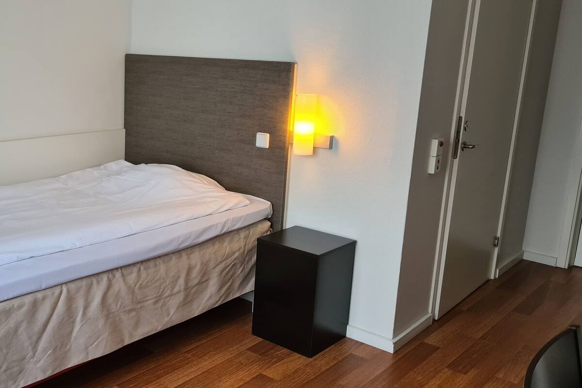 A single bed with a white mattress and a beige cover, a small black nightstand, a wall-mounted light, and a door with a handle.