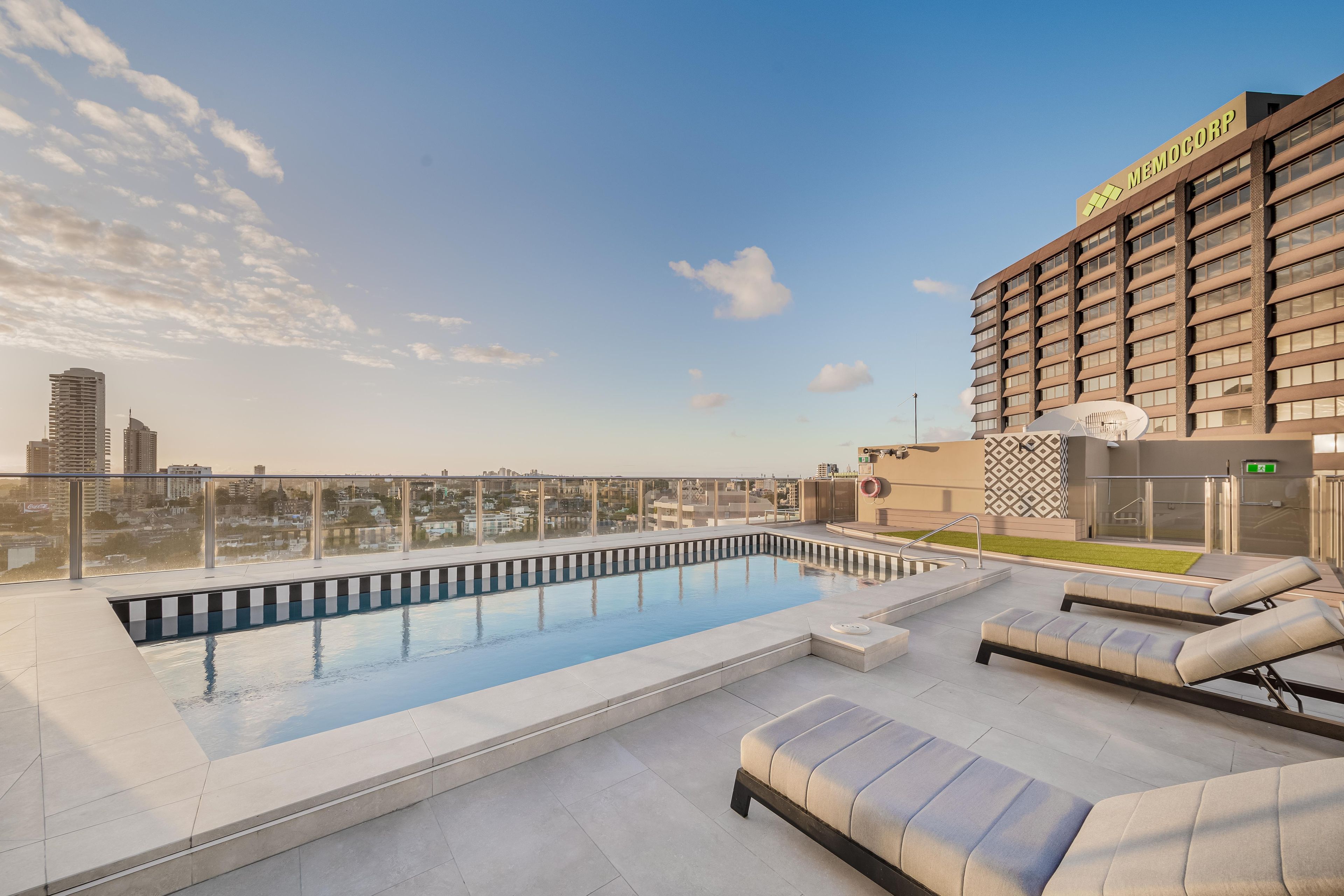 Un espace piscine sur le toit offrant une vue sur les gratte-ciel de la ville, un bâtiment arborant l’enseigne « MEMOCORP », et un salon avec des sièges.