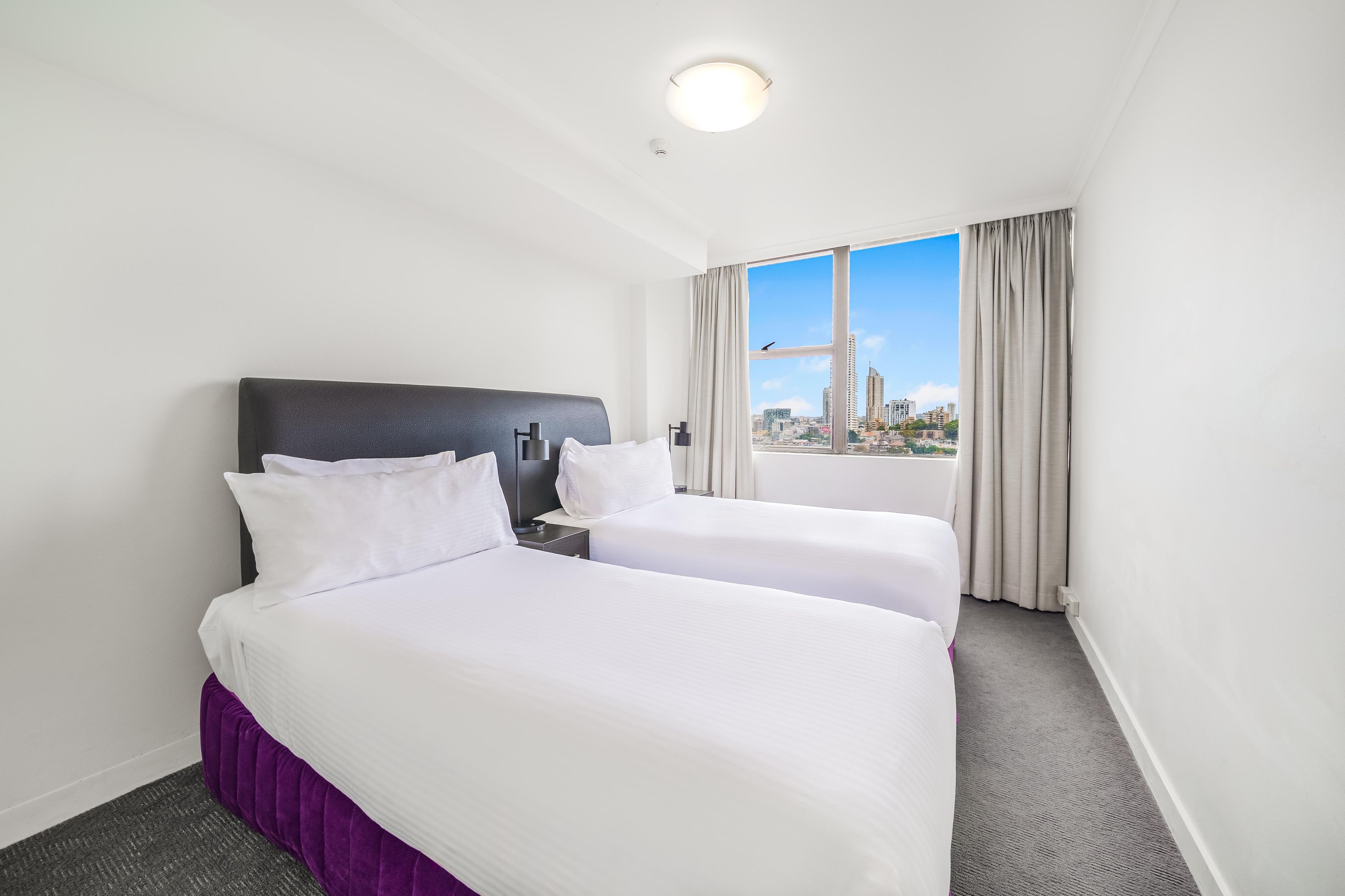 Habitación de hotel con dos camas, una ventana con cortinas y vista a la ciudad.