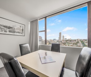 Een vergaderruimte met een tafel en vier stoelen, een foto van een stadsgezicht in een lijst aan de muur, en een uitzicht op een stadsgezicht door grote ramen.
