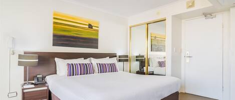 Ein Hotelzimmer mit einem großen Bett, Nachttischen, einer Lampe, einem Gemälde, einem Spiegel und einer Tür.