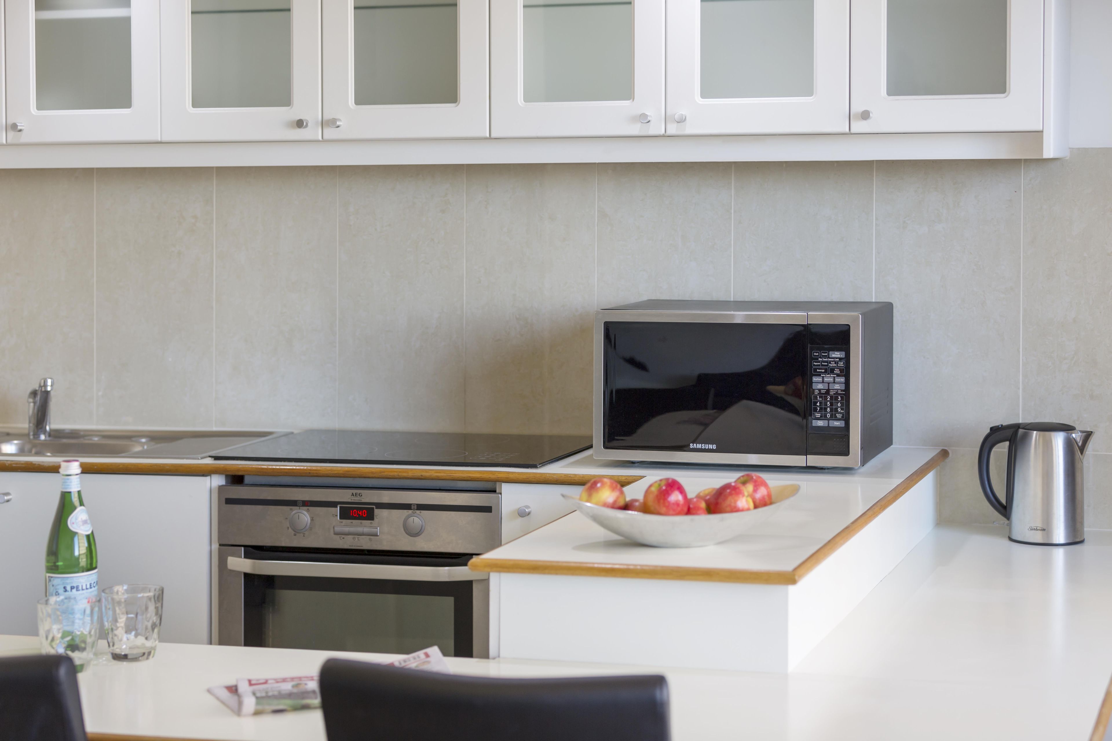 Una cocina con armarios blancos, un microondas, una tetera y un bol de manzanas sobre la encimera.
