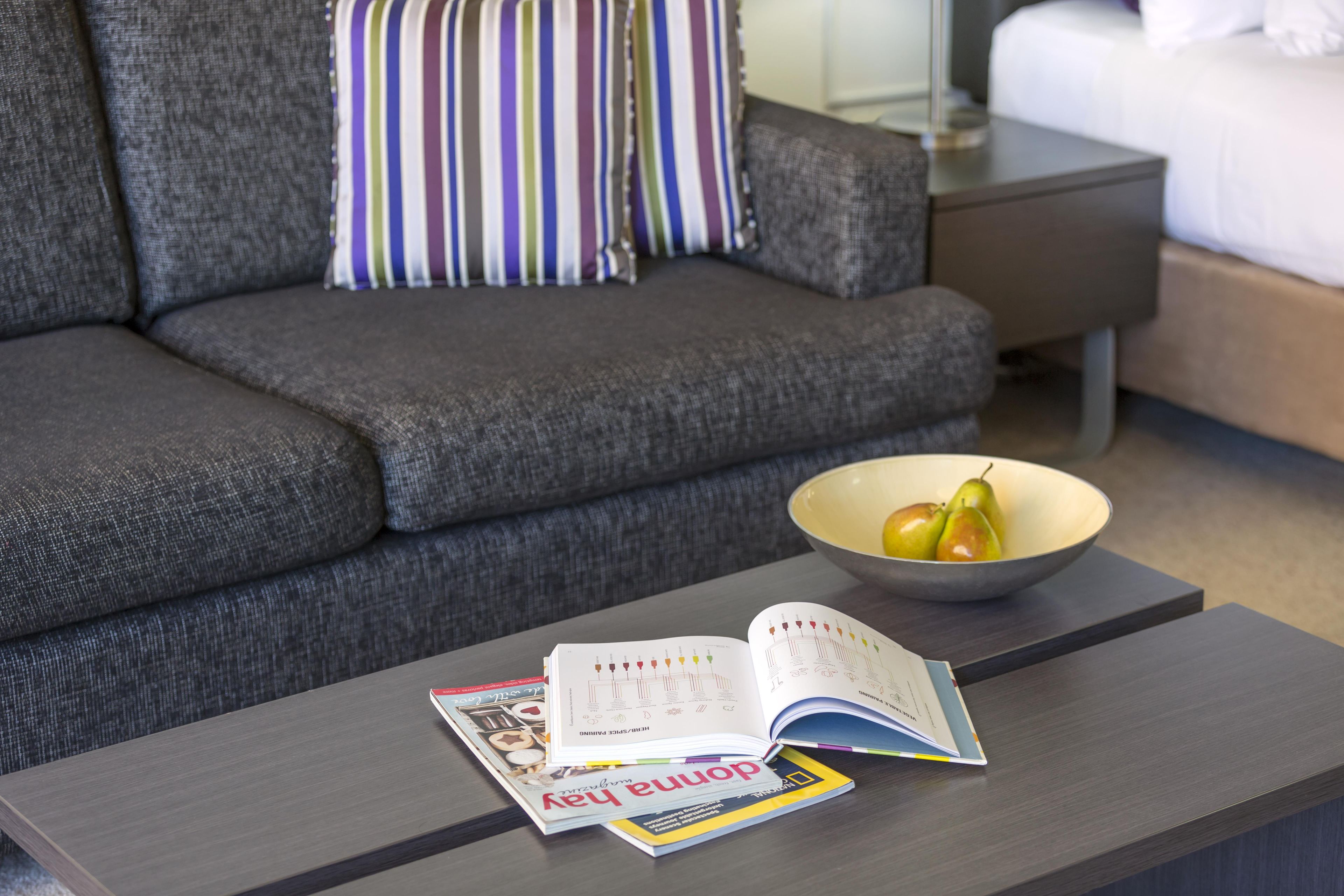 Ein modernes Wohnzimmer mit einem grauen Sofa, einem gestreiften Kissen, einem Nachttisch mit einer Lampe und einer Obstschale auf einem Couchtisch.
