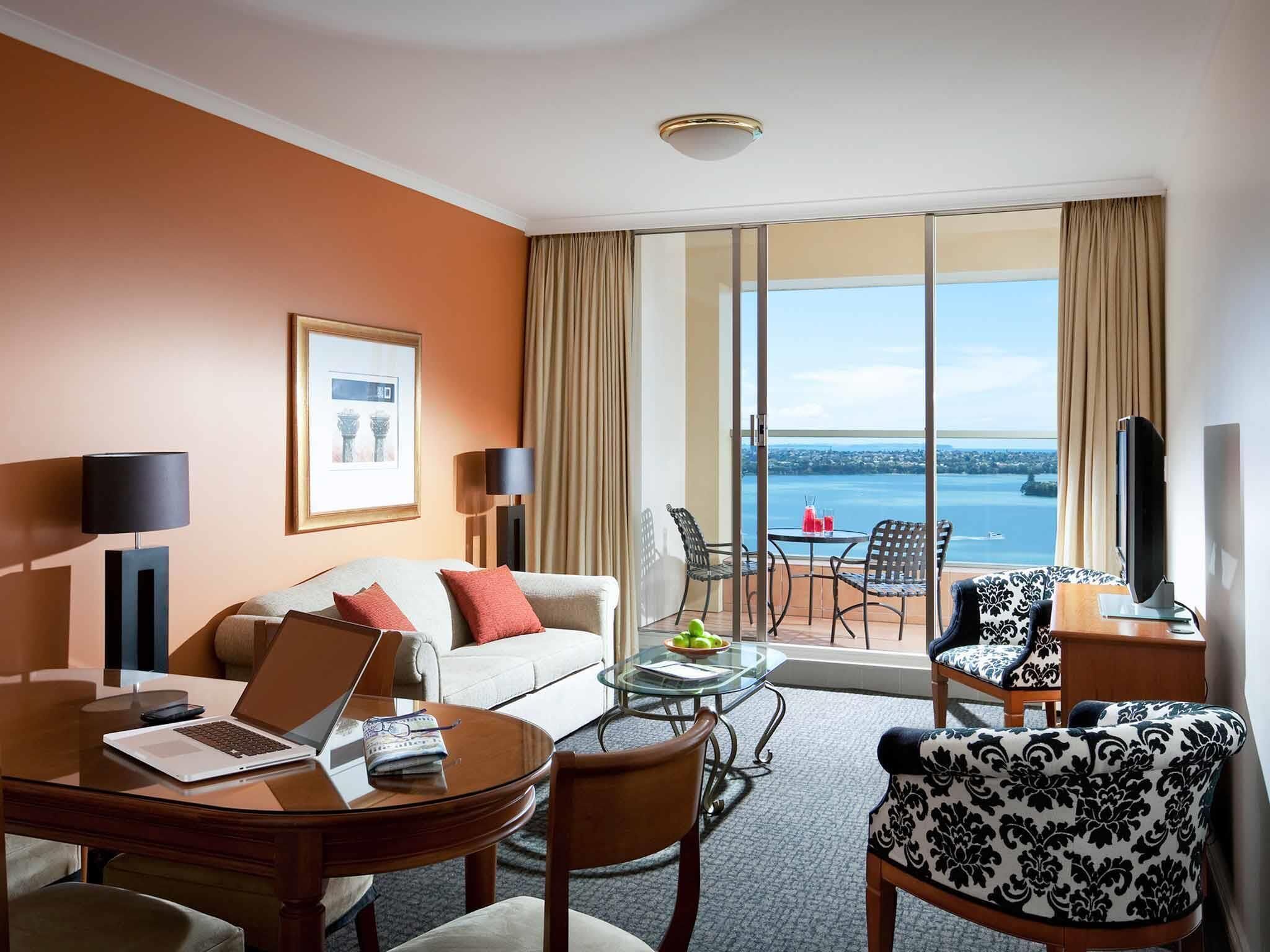 A hotel room with a sofa, a dining table with a laptop, a TV, and a balcony with a view of the sea.