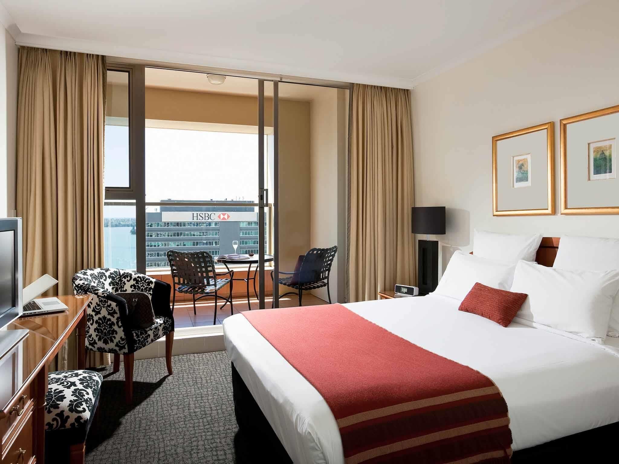 A hotel room with a bed, a desk with a computer, a chair, a TV, and a view of the cityscape.