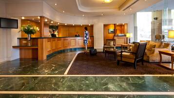 A hotel lobby with a reception desk, a large vase of flowers, a seating area with chairs and a sofa, and a marble floor.