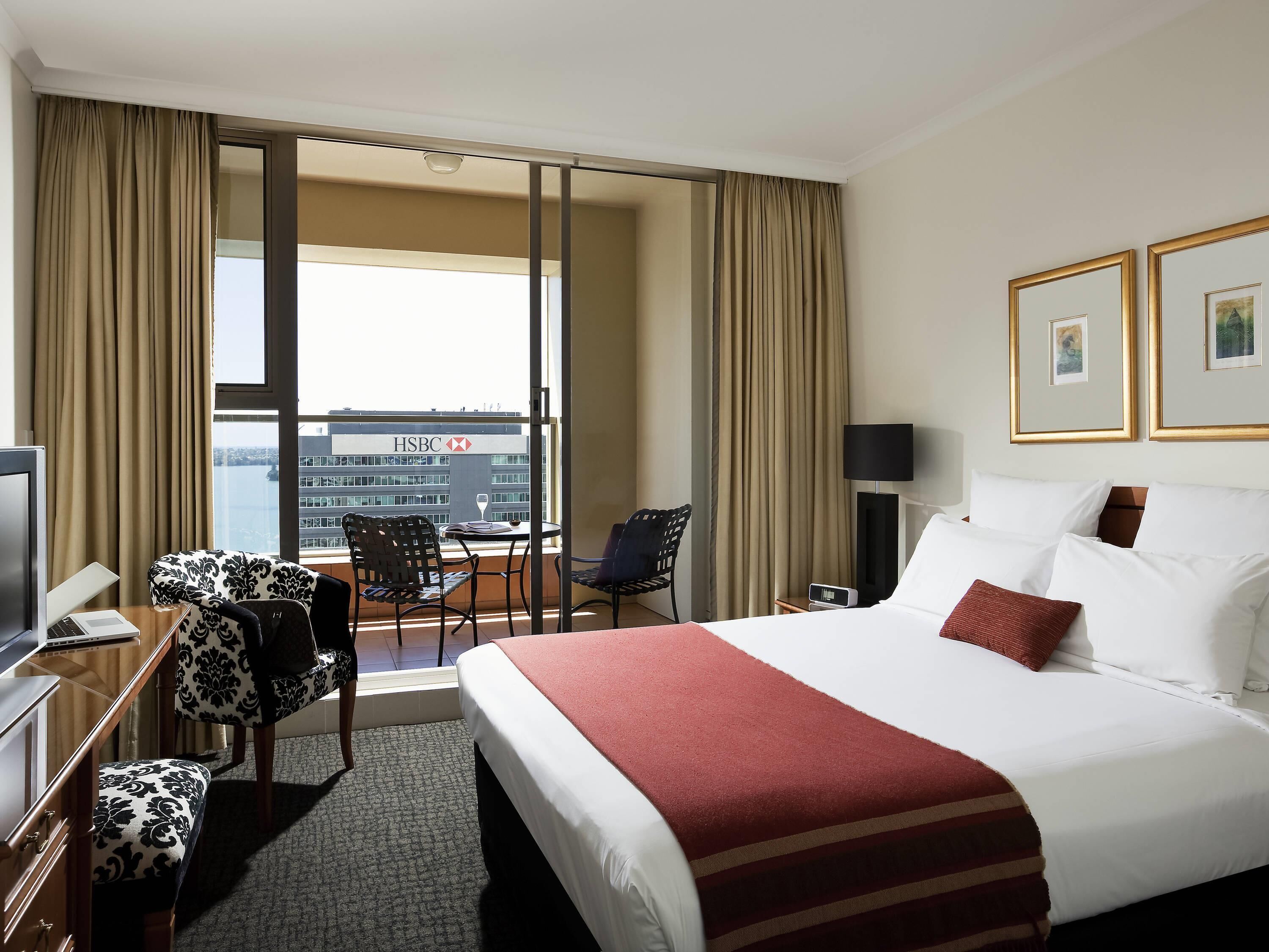 A hotel room with a bed, a desk with a computer, a chair, a table, a lamp, and a view of the HSBC building.