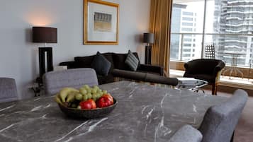 A modern hotel room with a marble dining table, a fruit bowl, a sofa, and a large window with a city view.