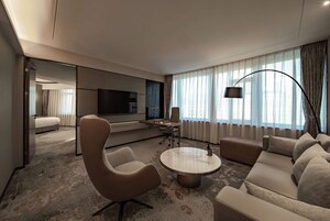 A modern hotel room with a sofa, armchair, coffee table, TV, and a large window with curtains.