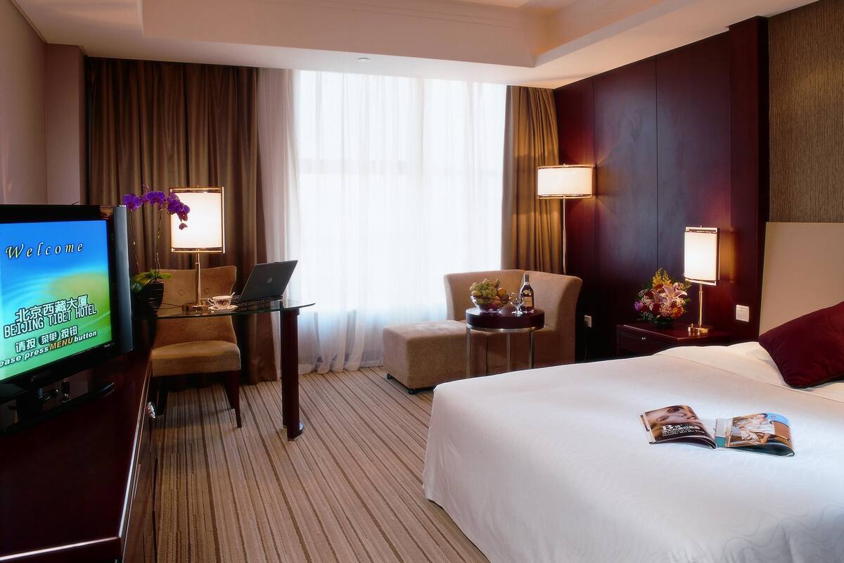 Une chambre d’hôtel comprenant un lit, un bureau avec un ordinateur portable, un téléviseur affichant un message de bienvenue, une chaise, une petite table avec un vase et une fenêtre avec des rideaux.