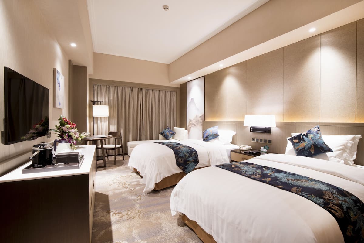 Habitación de hotel con dos camas, un escritorio con lámpara, televisor y una pequeña mesa con flores.