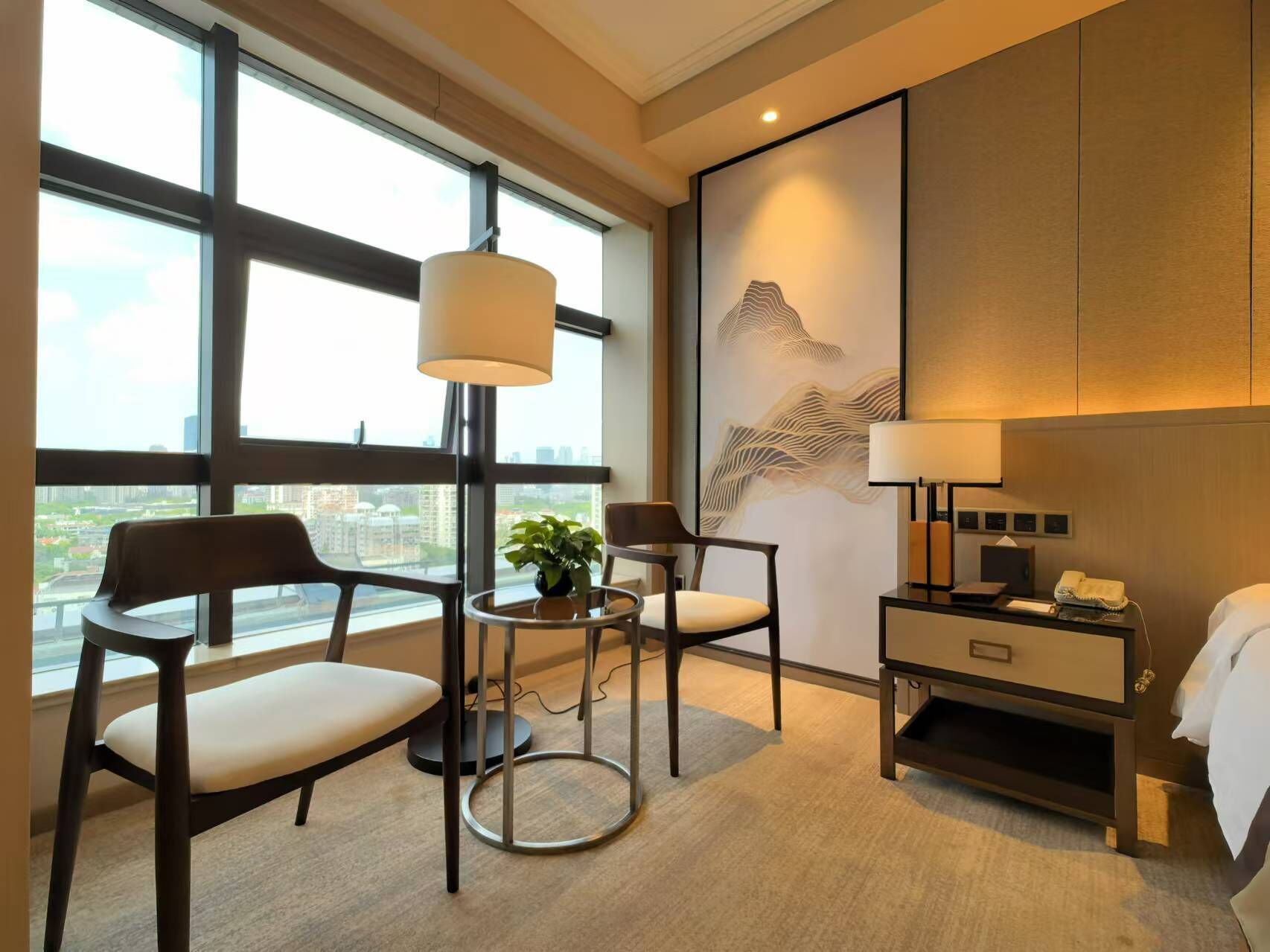 A hotel room with a large window, two chairs, a small table with a plant, a bedside table with a lamp, and a bed.