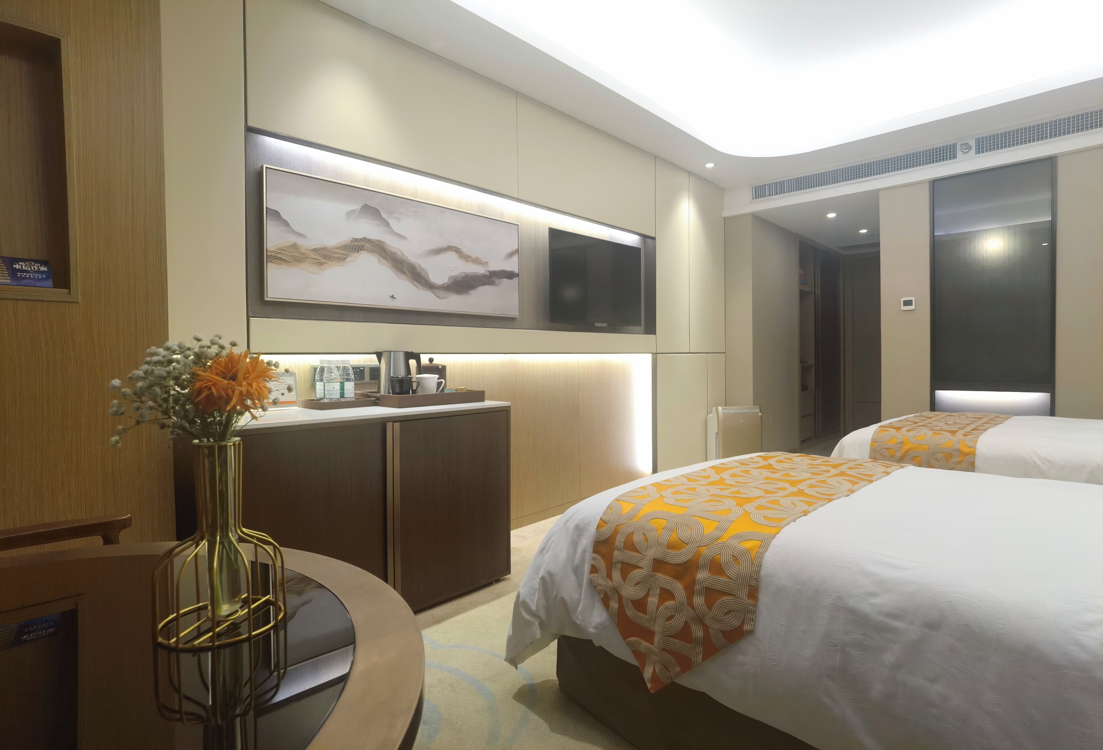 A hotel room with two beds, a desk, a coffee maker, a vase with flowers, and a mounted artwork.