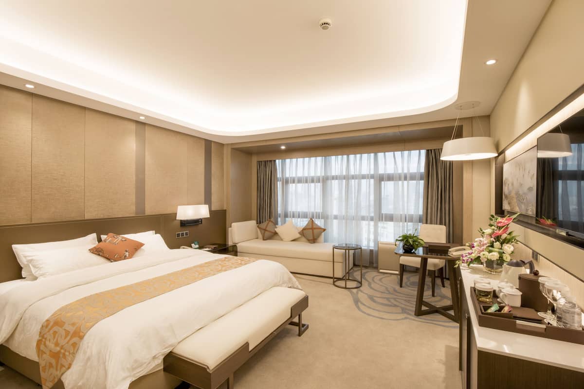 Una habitación de hotel moderna con una cama grande, un sofá, un escritorio con una silla y un ventanal con cortinas.