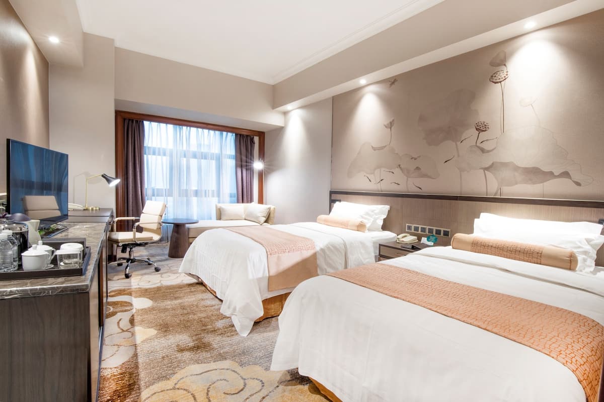 Habitación de hotel con dos camas, un escritorio con lámpara, televisión y una ventana grande con cortinas.
