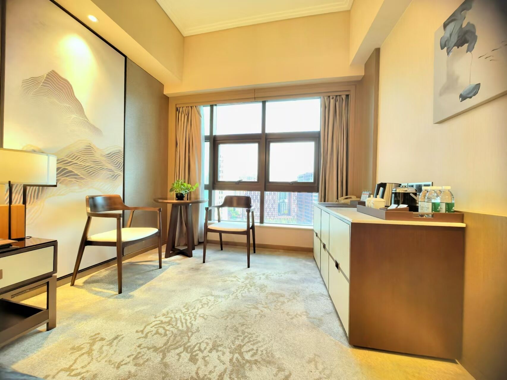 Habitación de hotel con un ventanal grande, un escritorio con teléfono y artículos de aseo, dos sillas y un adorno en la pared.