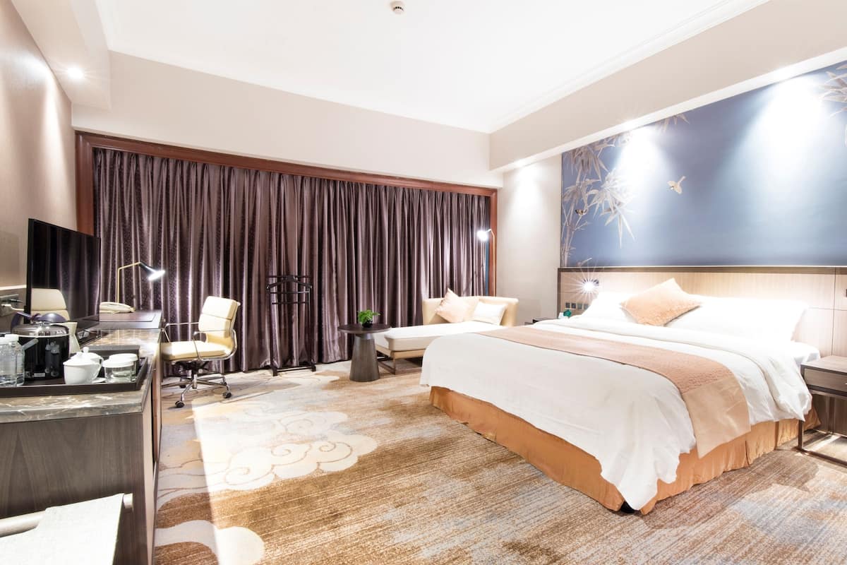 Una habitación de hotel moderna con una cama grande, un escritorio con computadora, una silla y un amplio ventanal con cortinas.