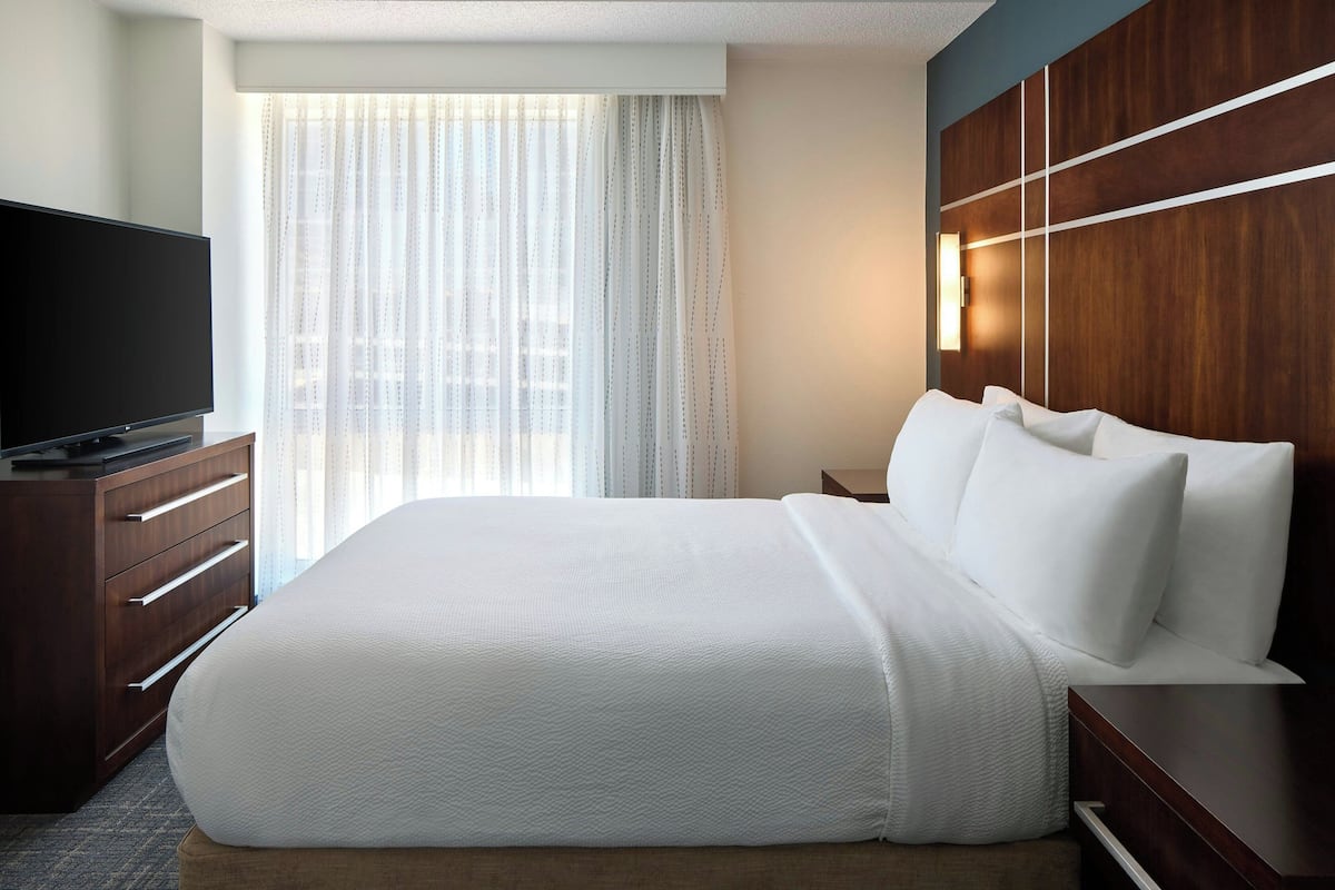 Ein Hotelzimmer mit einem großen Bett, einem Fernseher auf einer Holzkommode und einem Fenster mit durchsichtigen Vorhängen.