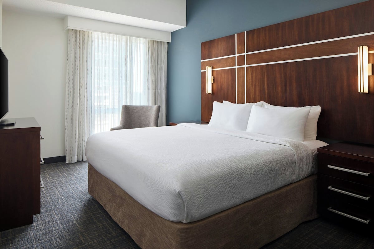 Ein Hotelzimmer mit einem großen Bett, einem Holz-Kopfteil, einem Schreibtisch mit Stuhl und einem Fenster mit Vorhängen.