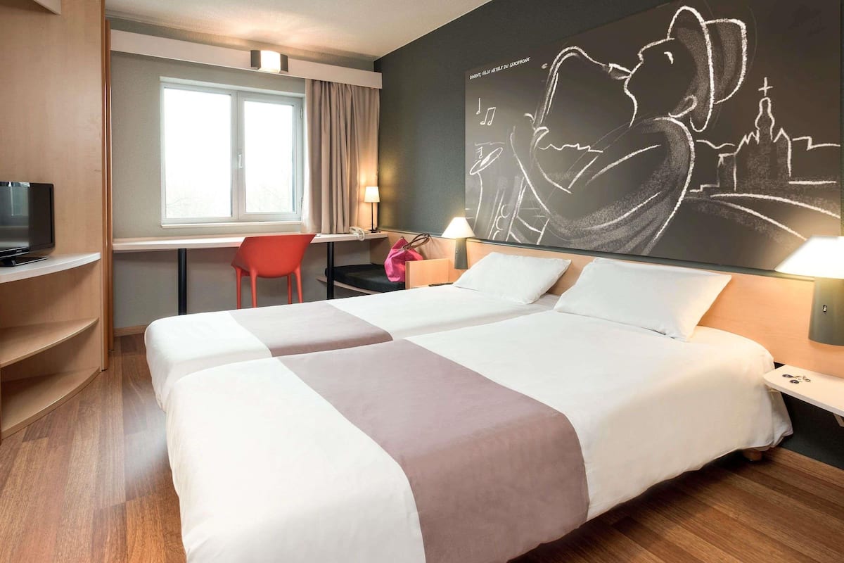 A hotel room with two beds, a desk, a chair, a TV, and a wall art piece.