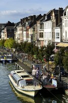 A canal-side town with a row of historic buildings, a docked boat, and people gathered on the promenade.