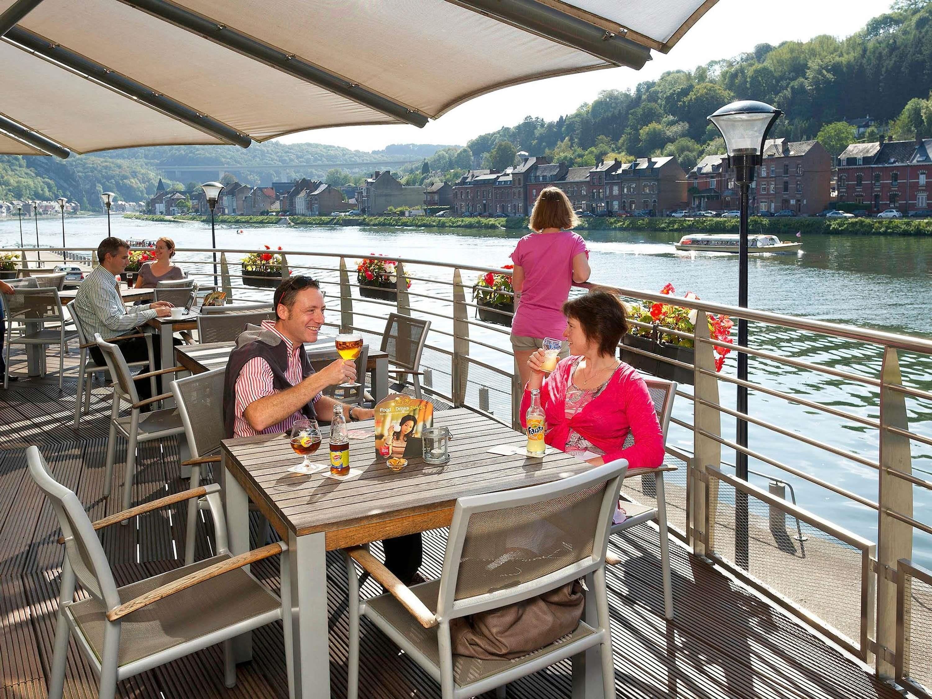 Un restaurante junto al río con terraza, mesas, sillas y vistas al río y edificios al fondo.