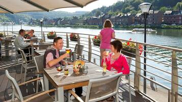 Un restaurante junto al río con terraza, mesas, sillas y vistas al río y edificios al fondo.