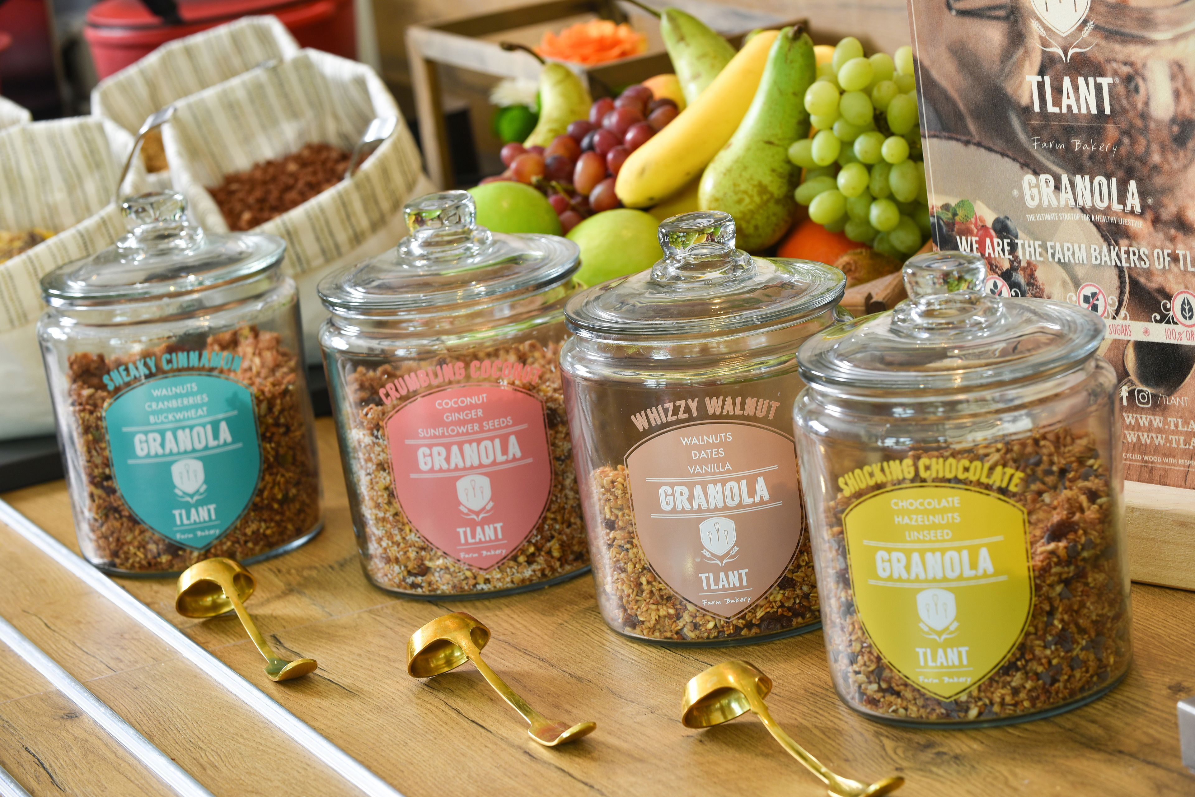 Cuatro frascos de granola con distintos sabores y una variedad de frutas y verduras al fondo.