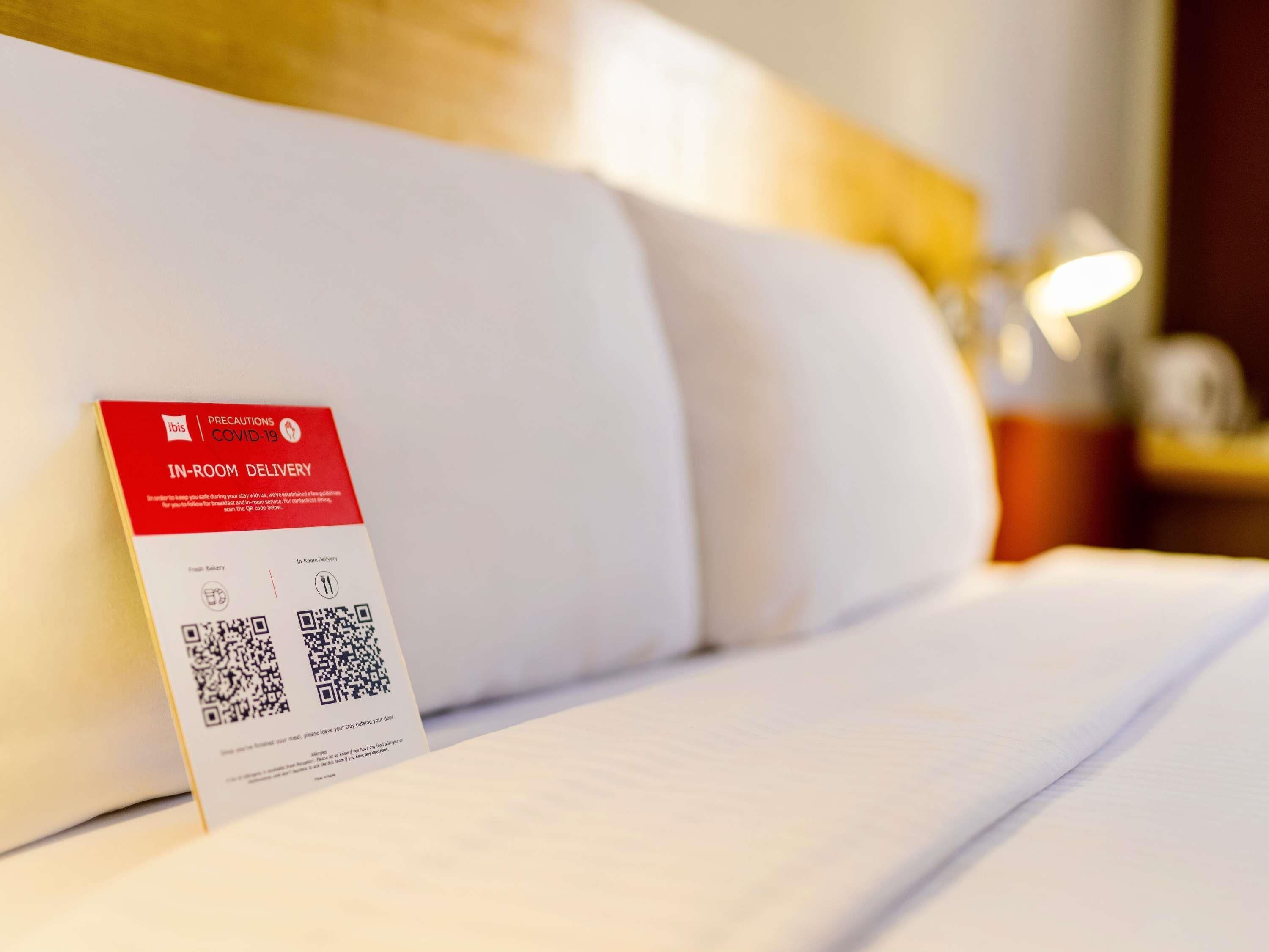 Una habitación de hotel con cama, almohadas, una mesita de noche y un cartel de servicio de entrega en la habitación.