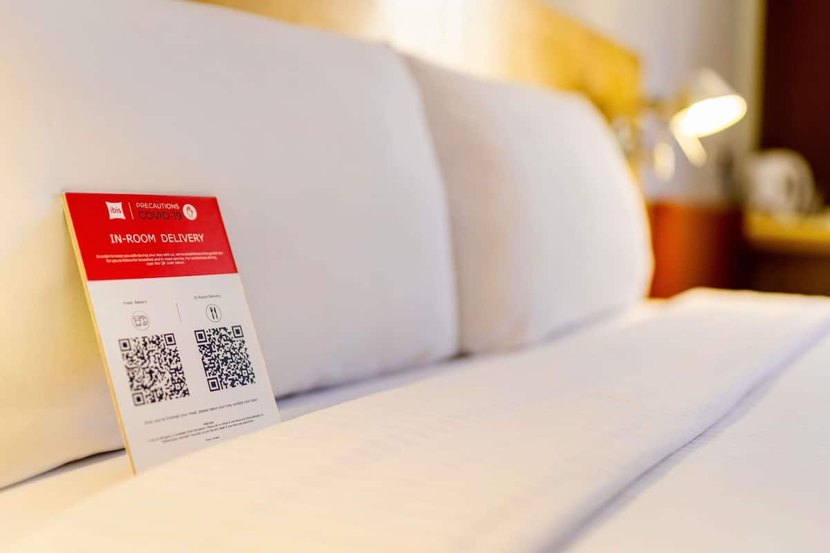 A hotel room with a bed, pillows, a bedside table, and a sign for in-room delivery services.