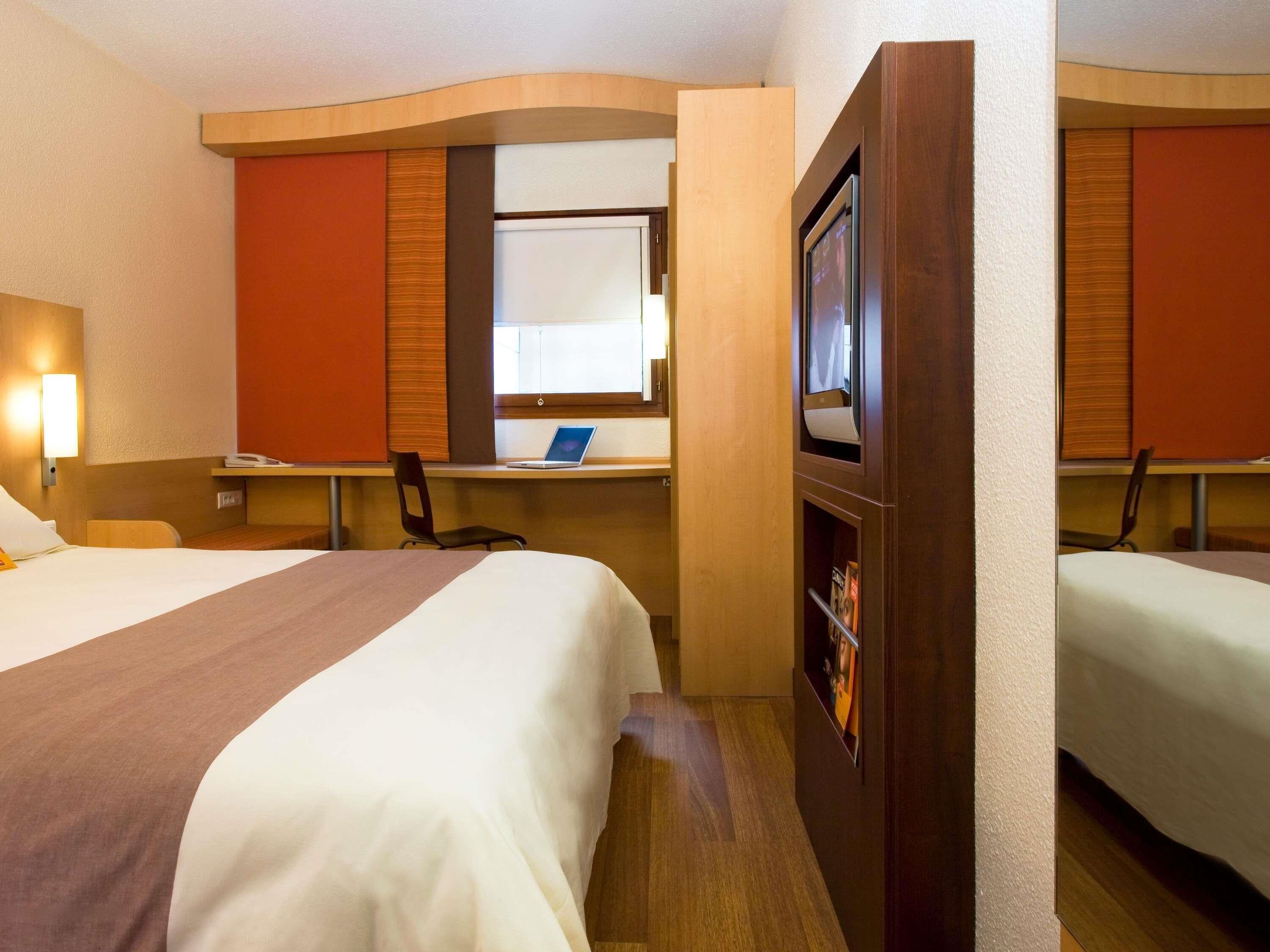 Een hotelkamer met twee bedden, een bureau met een laptop, een televisie en een raam.