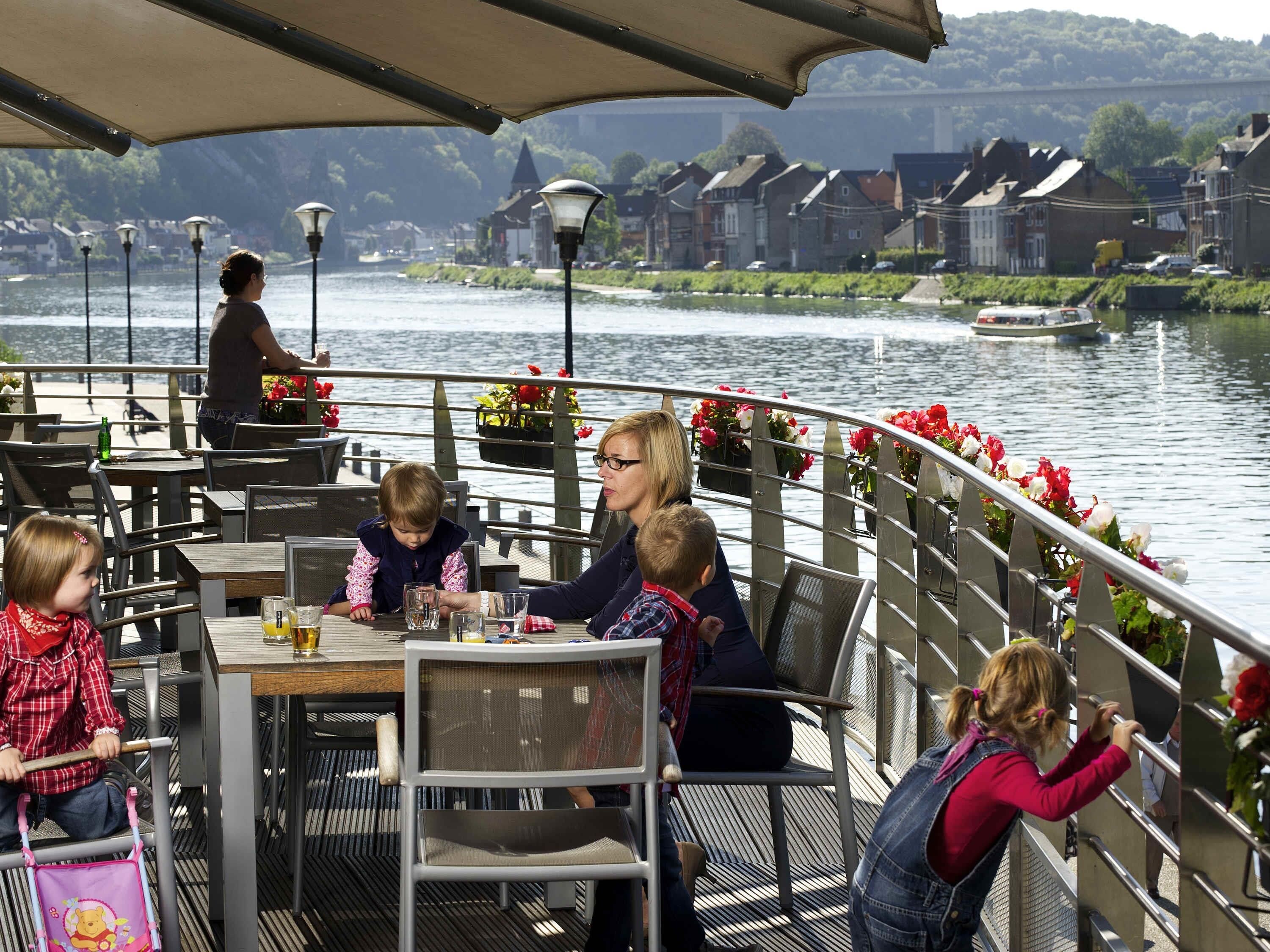 Un espace de restauration au bord de la rivière, avec des tables et des chaises, une rambarde ornée de fleurs et une vue sur la rivière et les bâtiments.