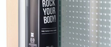 Una botella negra de gel de ducha con el texto “ROCK YOUR BODY!” y “Limpiador para cabello, cuerpo y rostro” en una pared de baño.