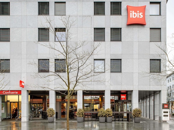 A building with multiple windows, a tree in front, and signs for 'Confiserie' and 'ibis' hotel. - ibis Baar Zug (Baar)
