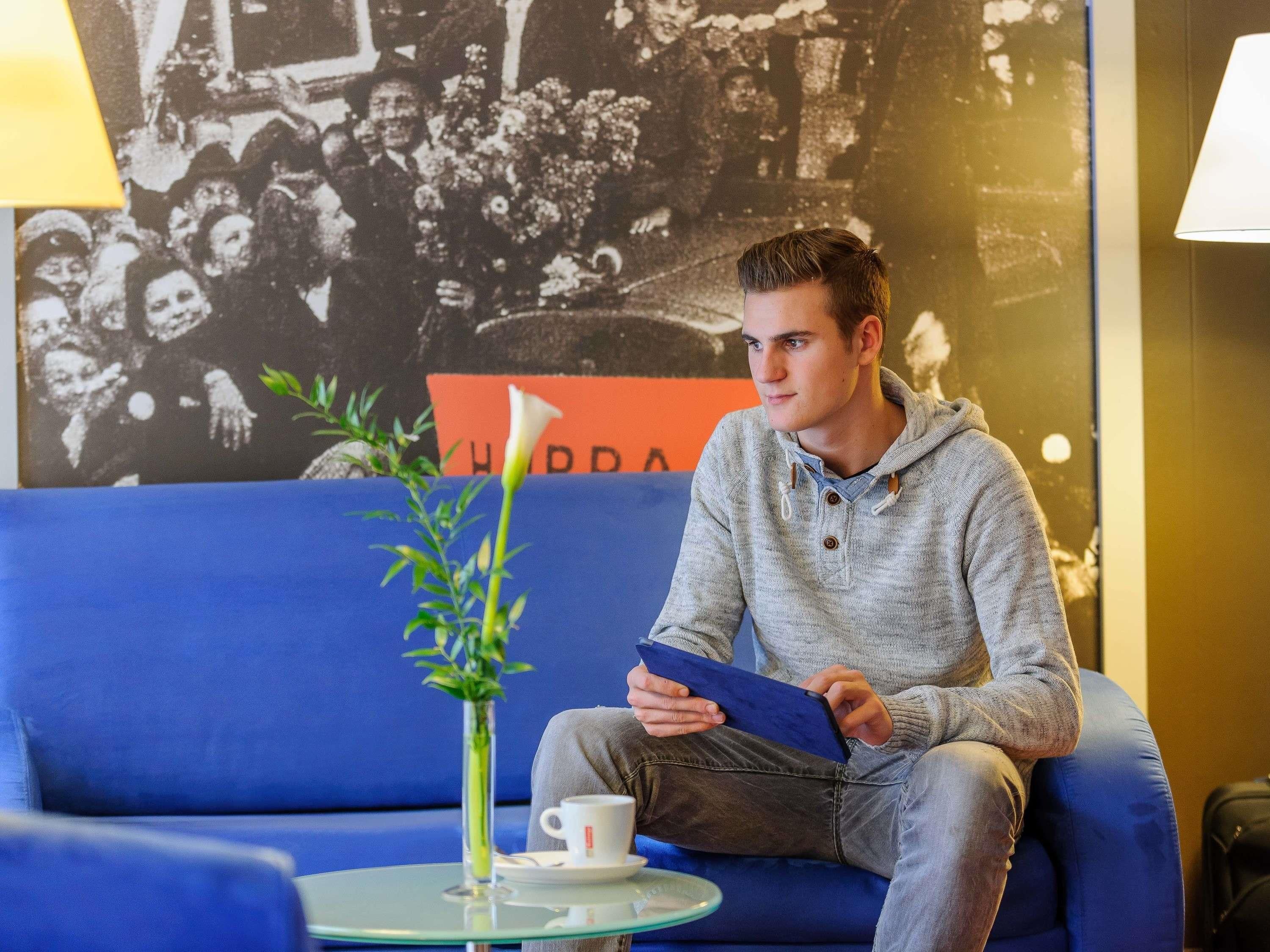 Ein Mann sitzt auf einem blauen Sofa, neben ihm ein Tablet, eine weiße Tasse und ein kleiner Tisch mit Pflanze und Vase.