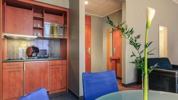 Habitación de hotel compacta con kitchenette, comedor con mesa de vidrio y zona de estar con sofá azul.
