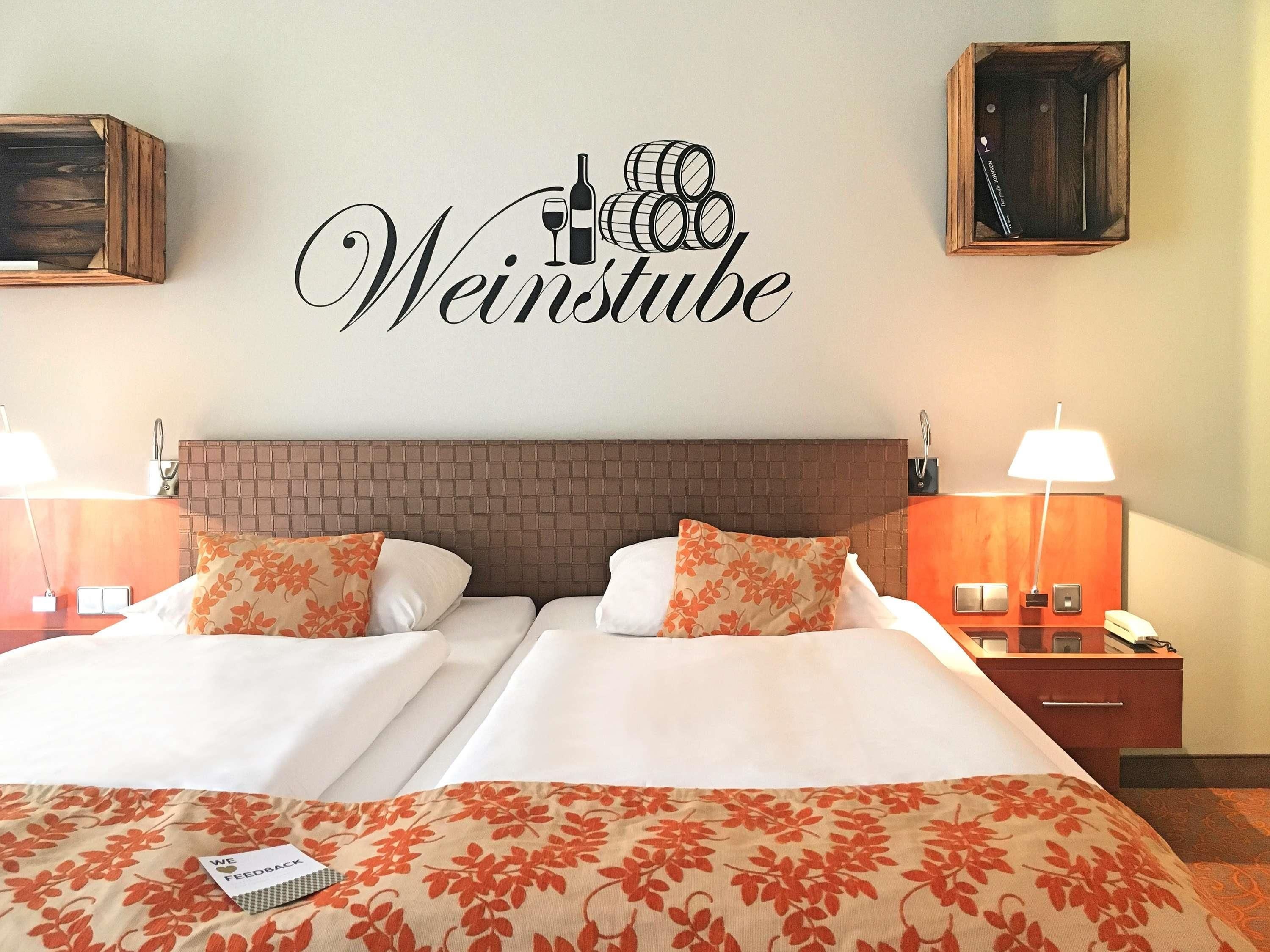 A hotel room with a double bed, two nightstands, a wall-mounted lamp, and a sign above the bed reading 'Weinstube'.