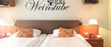 A hotel room with a double bed, two nightstands, a wall-mounted lamp, and a sign above the bed reading 'Weinstube'.
