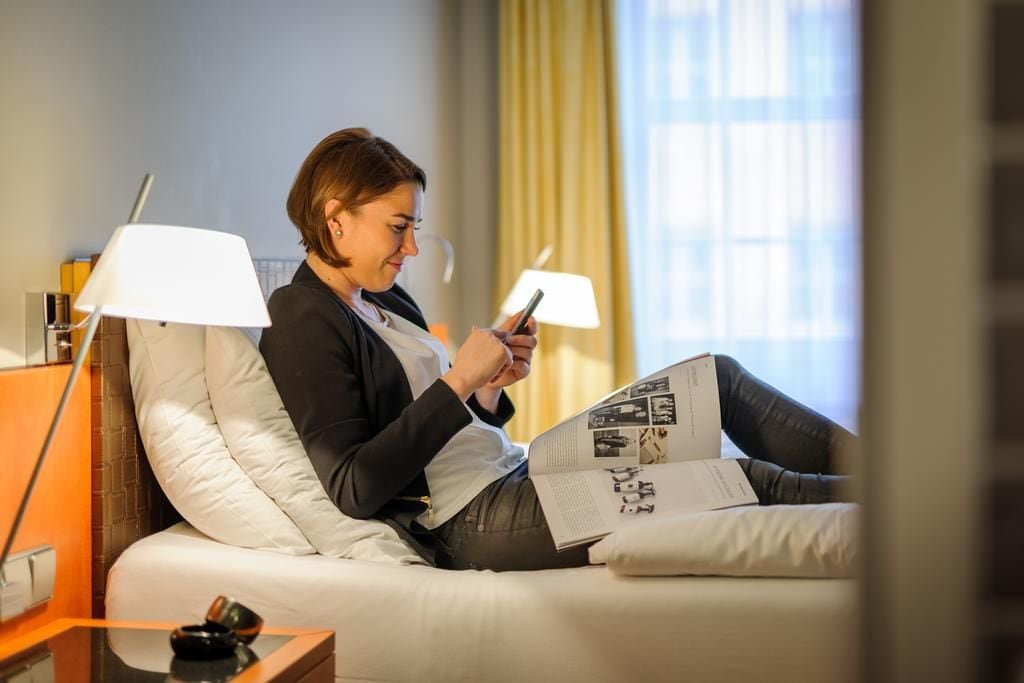 A person sitting on a bed with a book and a smartphone, a lamp on a bedside table, and a window with curtains.