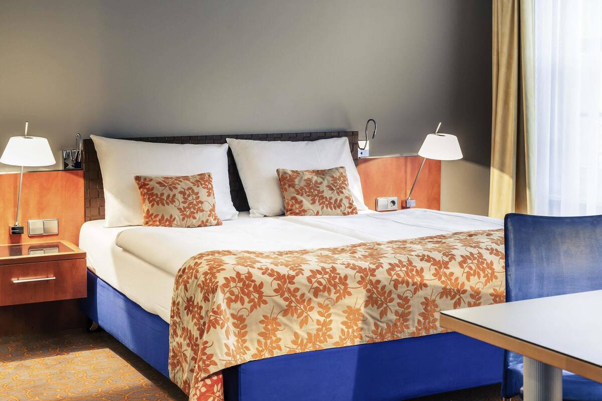 Hotellihuone, jossa on sänky, kaksi yöpöytää valaisimineen, työpöytä ja tuoli.