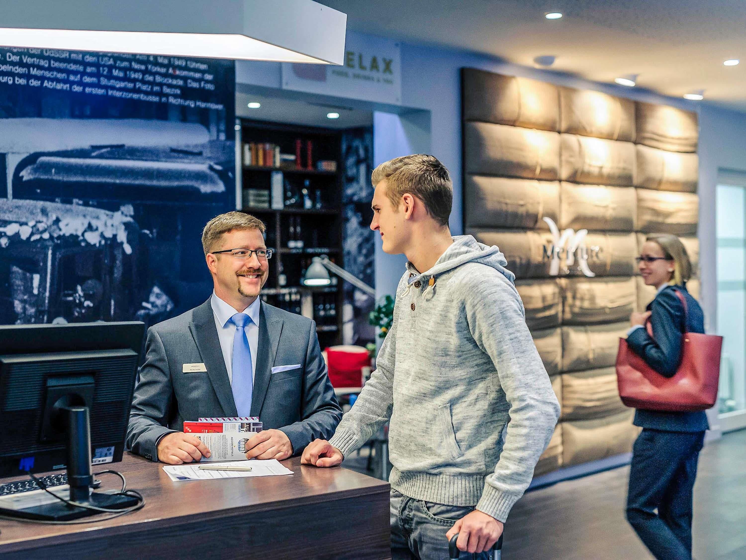 A reception desk with a man in a suit and a woman in a hoodie. There is a large wall with a logo and a woman carrying a red bag in the background.